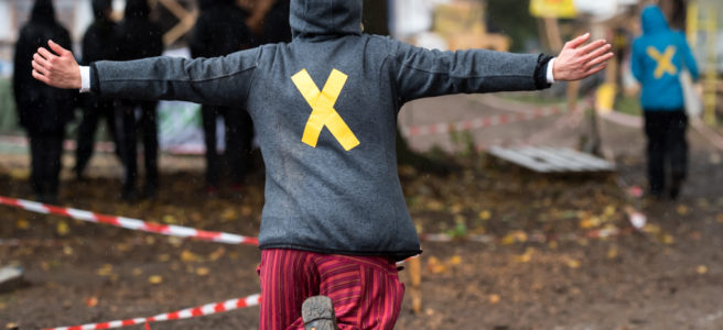 Foto eines Menschen von hinten, wie er mit ausgebreiteten Armen einen matschigen Weg entlang rennt, auf der Jacke ist ein gelbes X aufgeklebt, das Symbol des Bündnisses Alle Dörfer Bleiben