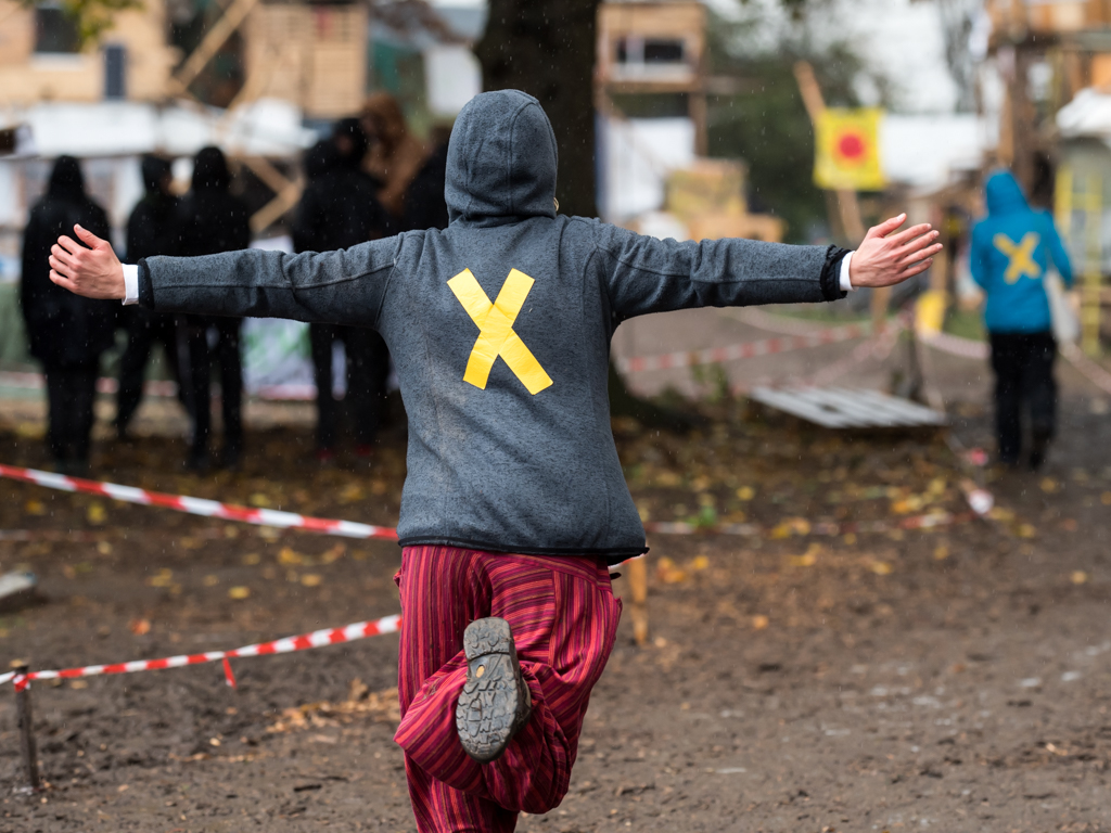 Foto eines Menschen von hinten, wie er mit ausgebreiteten Armen einen matschigen Weg entlang rennt, auf der Jacke ist ein gelbes X aufgeklebt, das Symbol des Bündnisses Alle Dörfer Bleiben